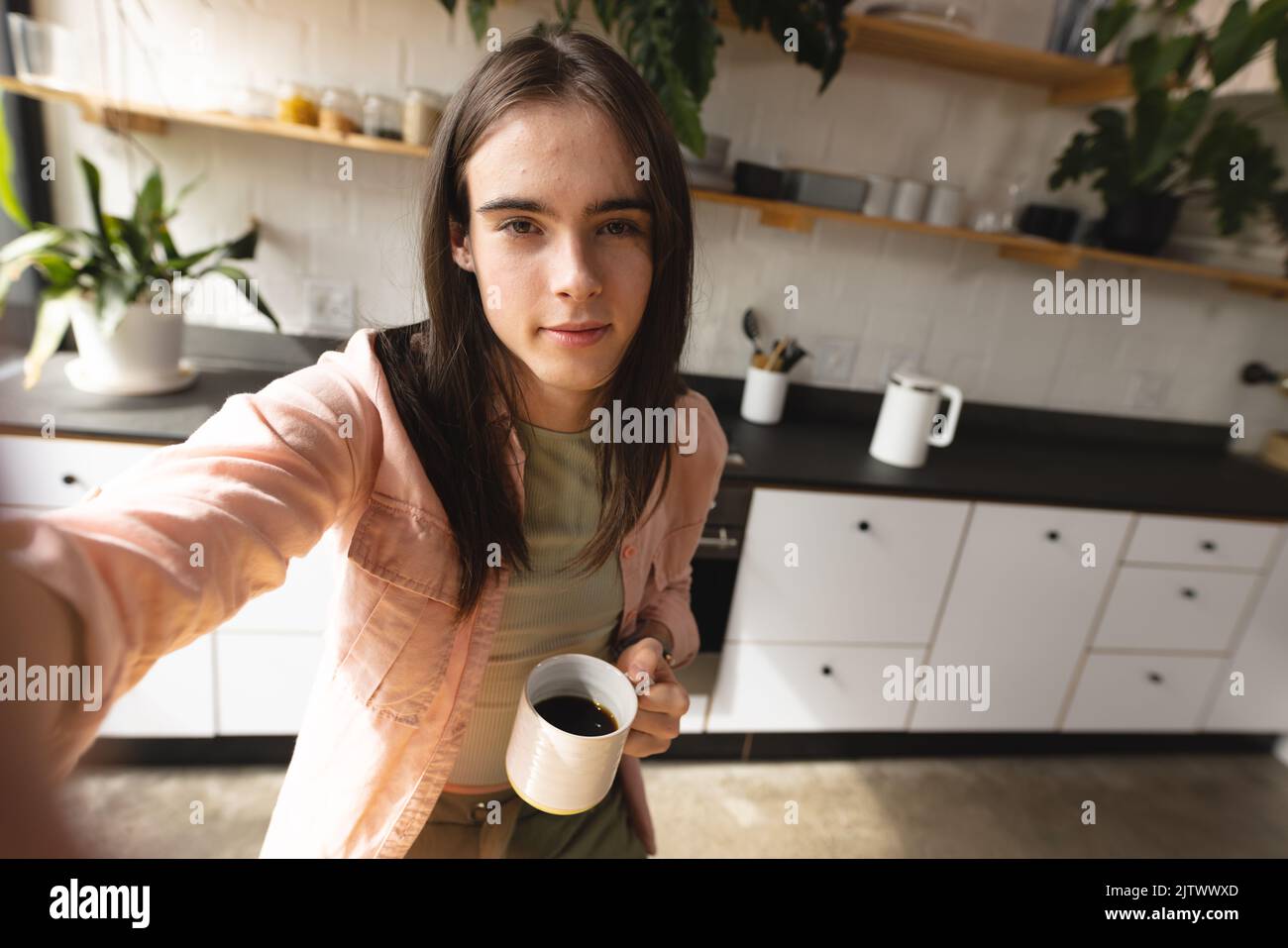 Porträt einer nicht-binären Transfrau, die eine Kaffeetasse in der Hand hält und die Kamera in der Küche zu Hause anschaut Stockfoto