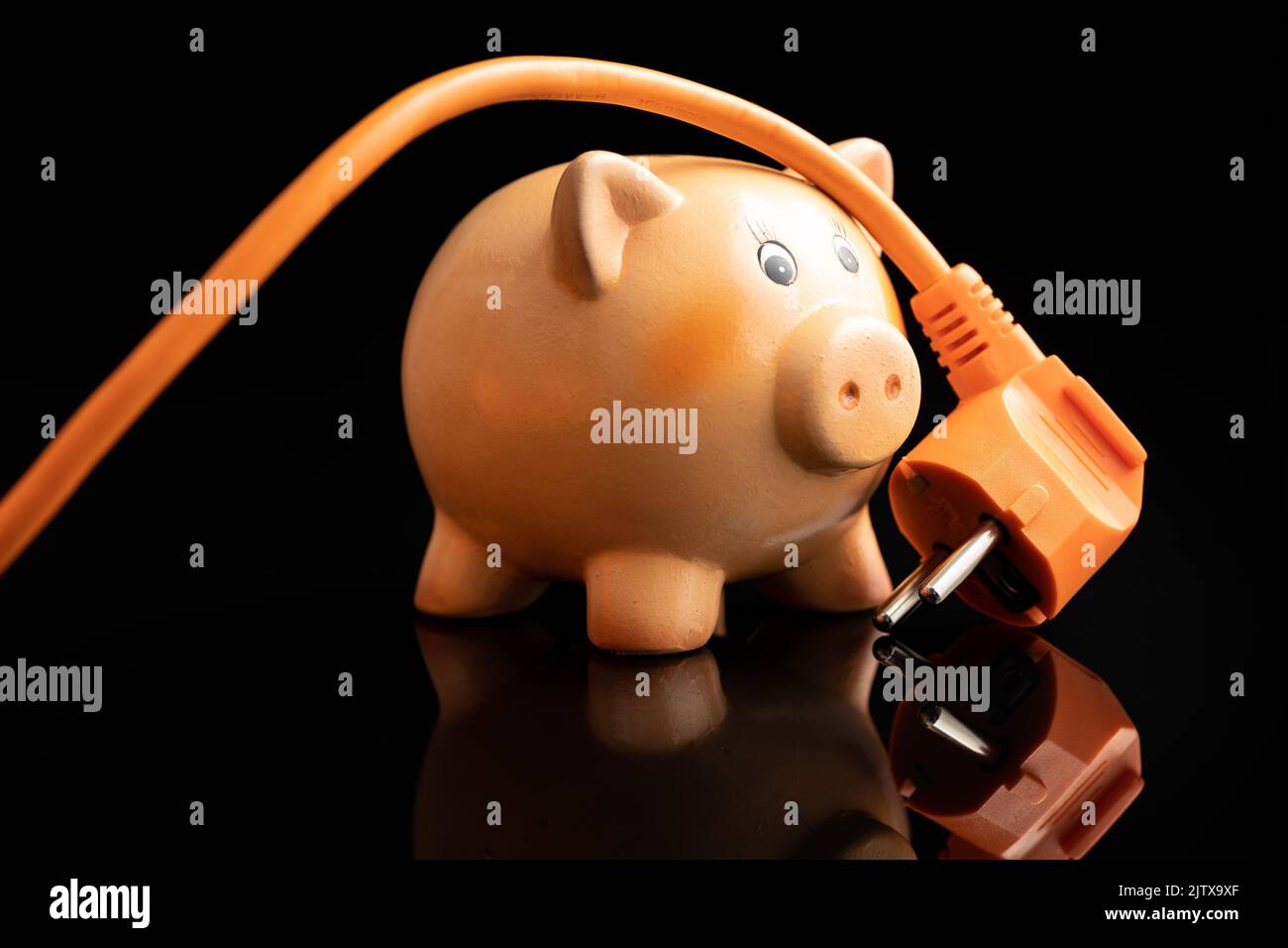 Orangefarbener Stecker und Sparschwein auf schwarzem Hintergrund. Konzept der steigenden Strompreise. Stockfoto