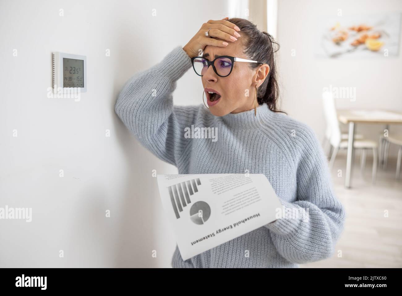 Die Frau kann ihren eigenen Augen die Summe auf ihrer Energierechnung nicht glauben und sieht schockiert über den Thermostat an der Wand neben ihr aus. Stockfoto
