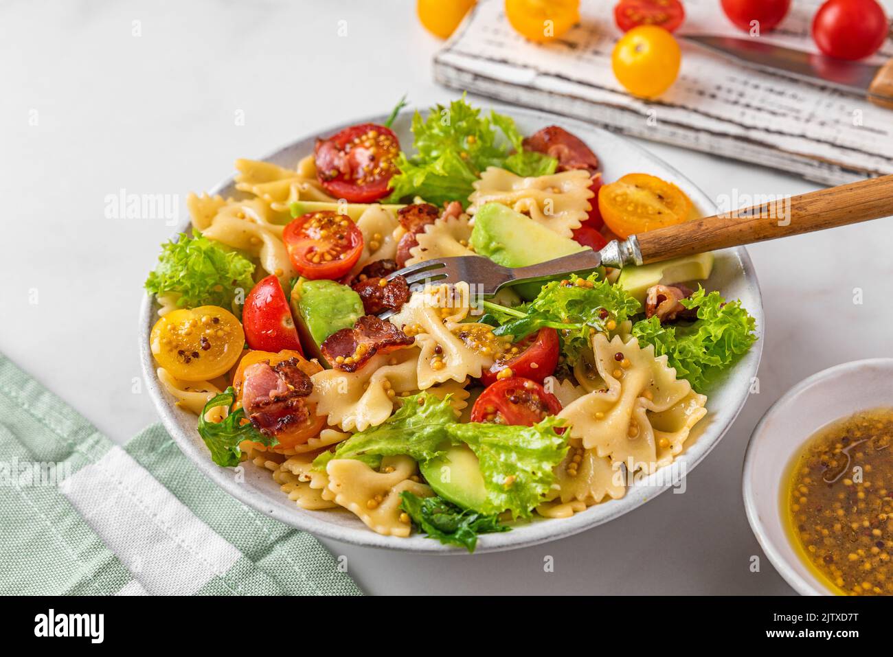 Kalter Sommer-Nudelsalat mit Speck, Tomaten, Avocado und Senf auf einem Teller mit Gabel auf weißem Hintergrund. Gesunde Ernährung Stockfoto