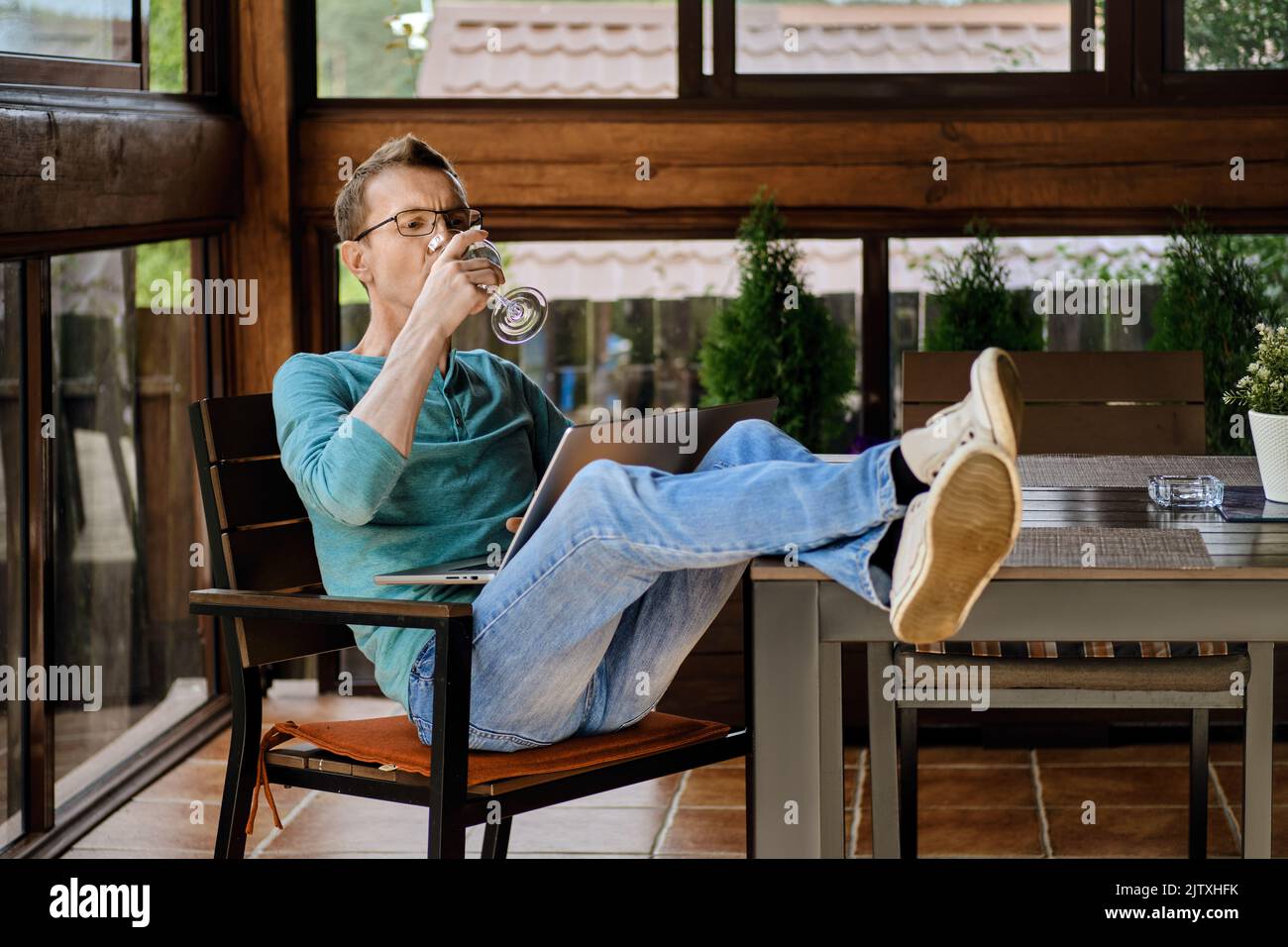 Mann mittleren Alters trinkt Wein, während er die Online-Übertragung auf dem Laptop auf der Veranda seines Landhauses sieht Stockfoto