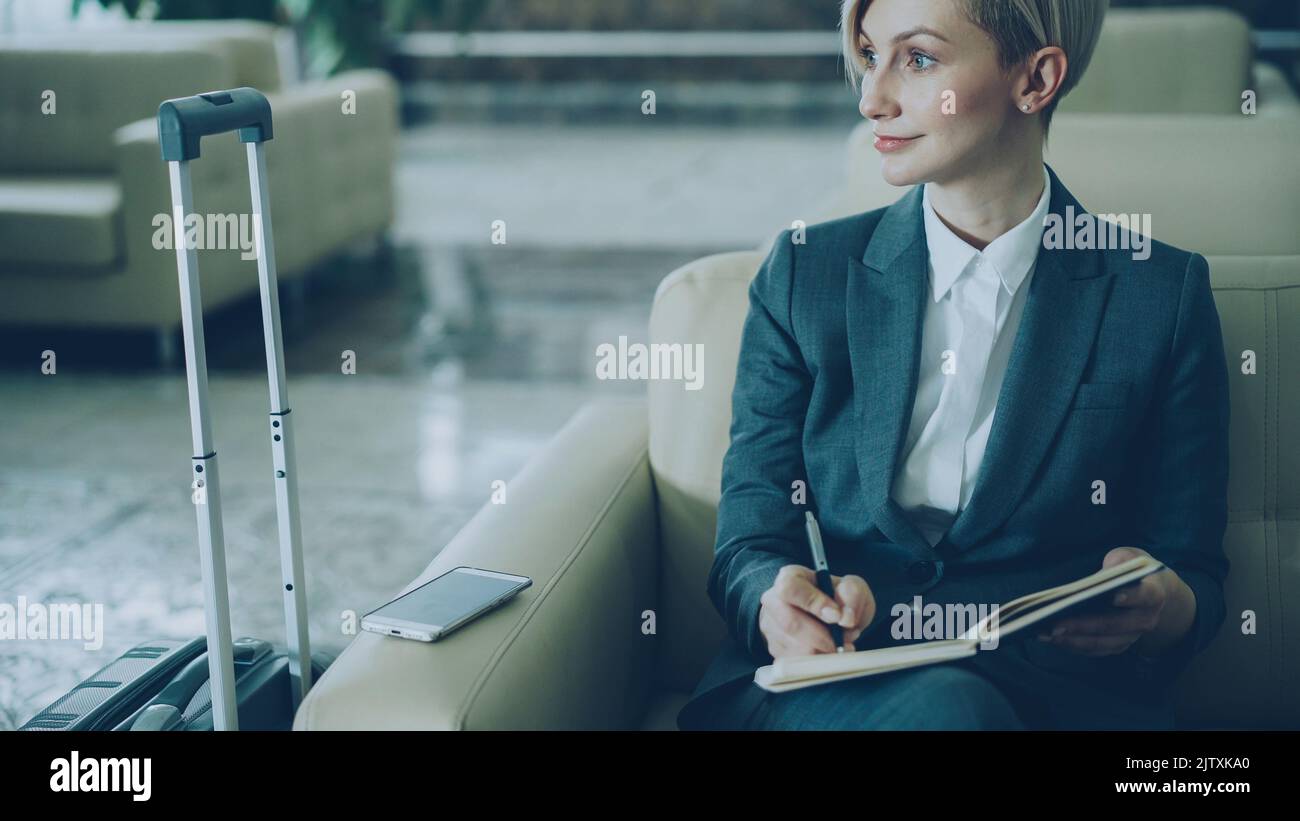 Attraktive blonde Geschäftsfrau, die in einem Sessel in der Hotellobby sitzt und lächelnd auf einem Notizblock schreibt und zur Seite schaut. Business-, Reise- und Mitarbeiterkonzept Stockfoto