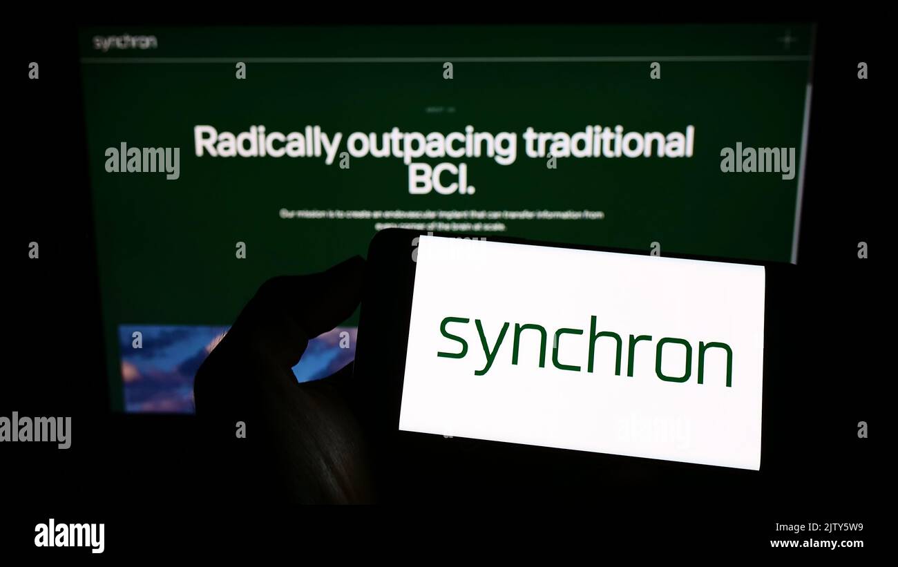 Person, die ein Mobiltelefon mit dem Logo des US-amerikanischen Unternehmens Synchron Inc. Auf dem Bildschirm vor der Unternehmenswebseite hält. Konzentrieren Sie sich auf die Telefonanzeige. Stockfoto