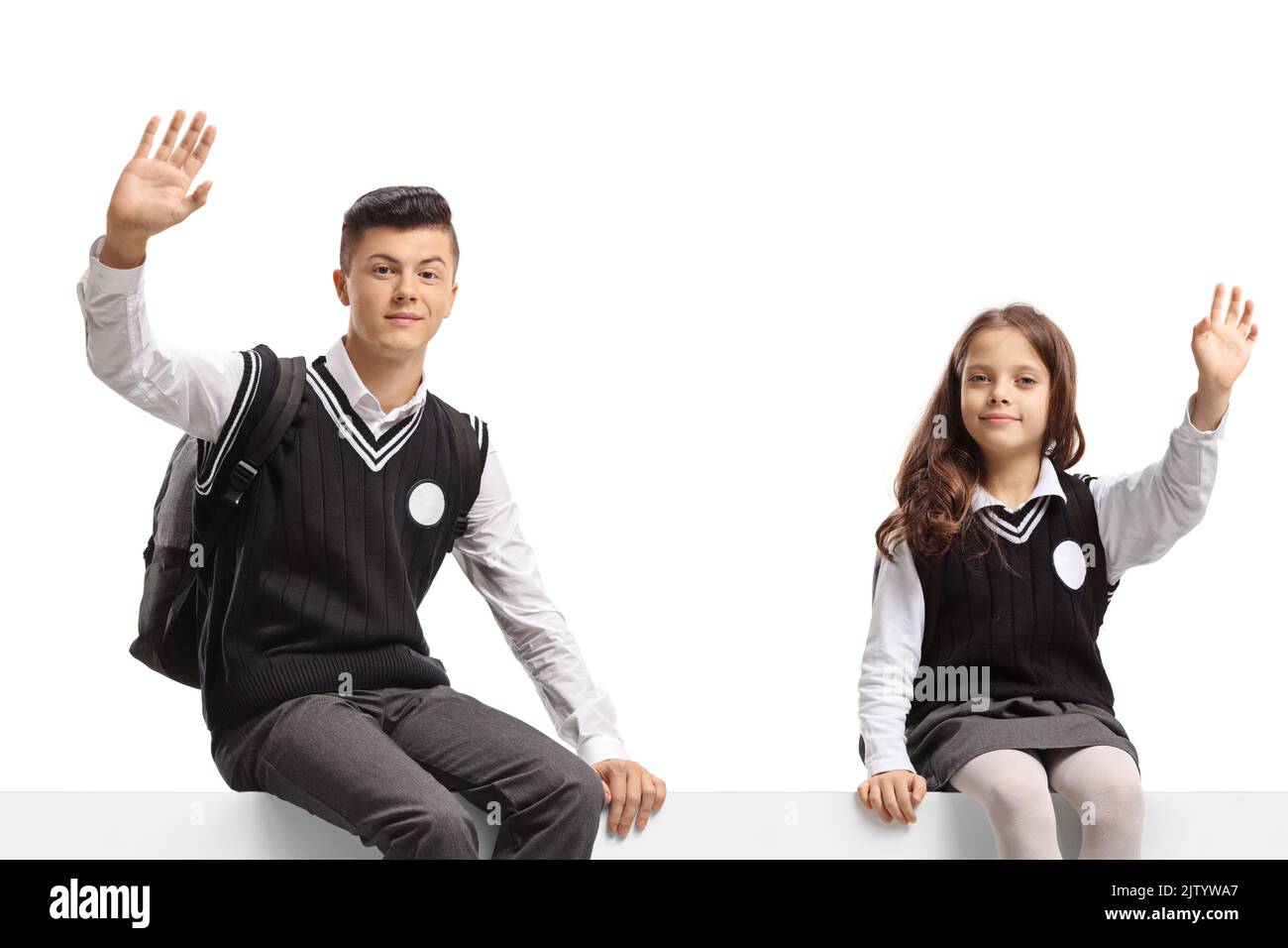 Männlicher Student und ein kleines Schulmädchen sitzen auf einer leeren Tafel und winken isoliert auf weißem Hintergrund auf die Kamera Stockfoto