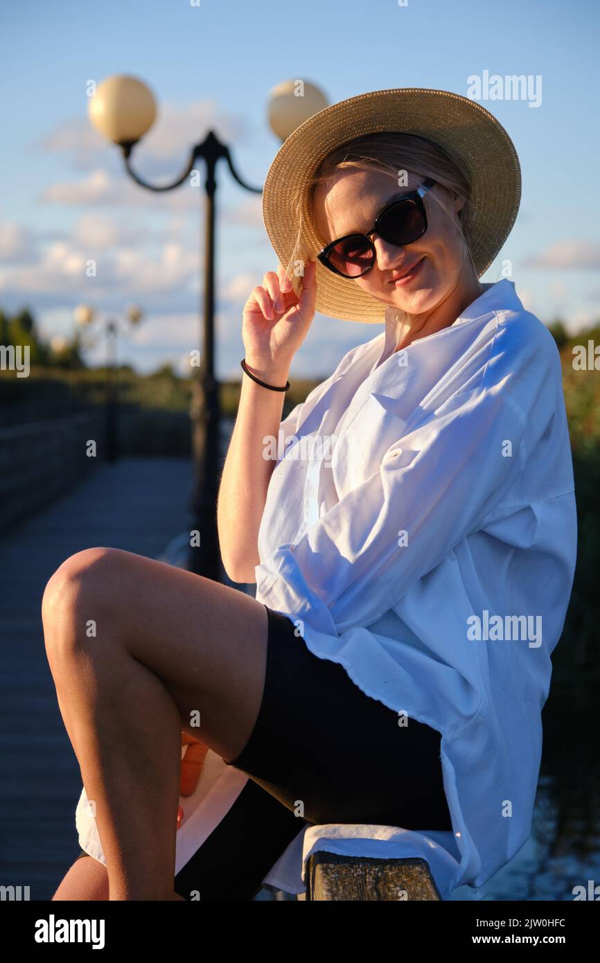 Junge glückliche blonde Frau in Strohhut und Sonnenbrille im Sommer Sonnenuntergang Park Blick auf die Kamera und lächelnd Stockfoto