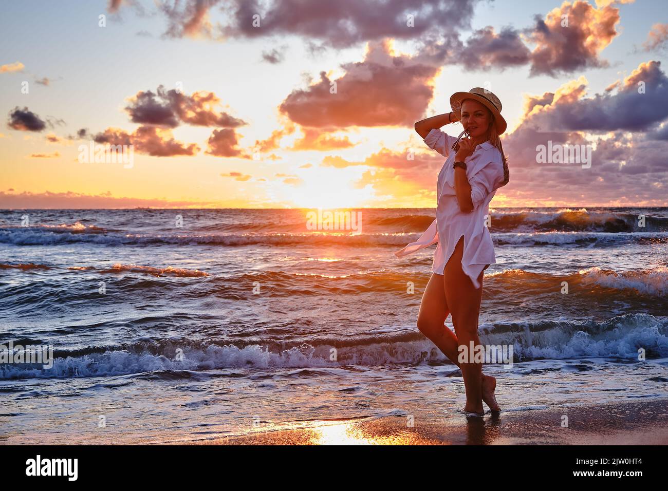 Sorglose Frau im Strohhut posiert im Sonnenuntergang am Meeresstrand. Vitalität gesundes Wohnkonzept. Mädchen im Pareo bei Sonnenuntergang auf stürmischem Meer. Stockfoto