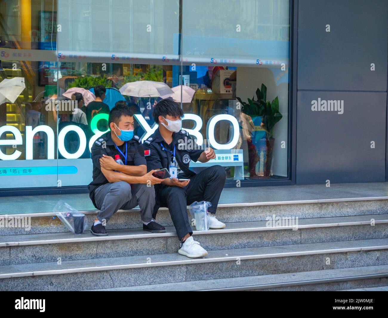 Zwei junge chinesische Männer sitzen auf den Stufen eines Gebäudeeingangs. Sie tragen Uniformen und schützende Gesichtsmasken. Stockfoto