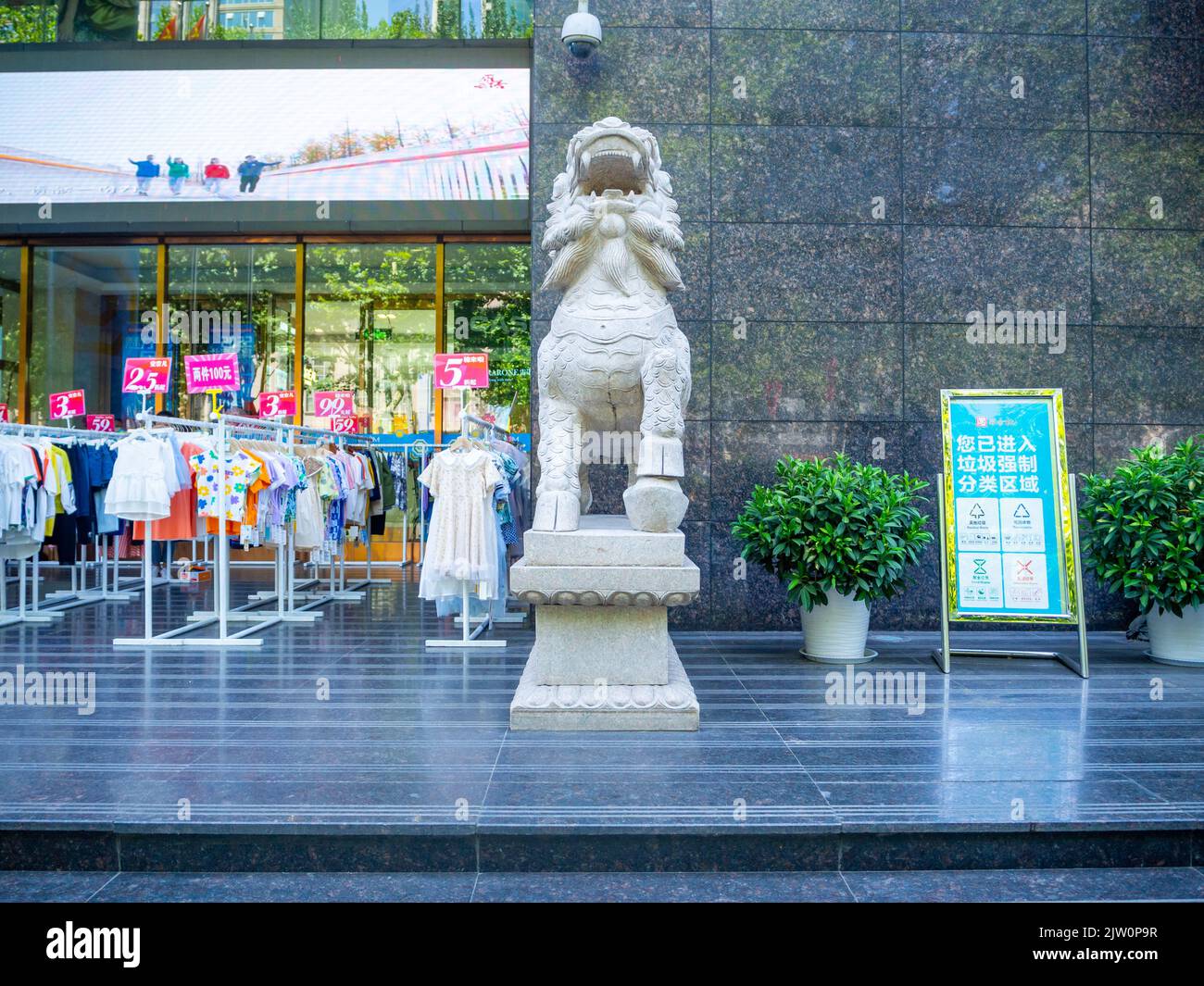 Traditionelle chinesische Skulptur in Form eines Löwen. Die Statue befindet sich vor einem Einzelhandelsgeschäft, in dem Kleidungsstücke im Außenbereich ausgestellt sind. Stockfoto