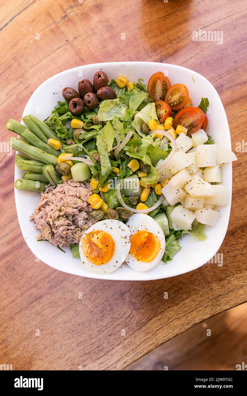 Thunfischsalat, Ei, Kartoffel, Salat, Zwiebel, Tomaten, Mais, schwarze Oliven und Spargel. Serviert in einer weißen Schüssel auf einem Holztisch. Gesunde Mahlzeit. Stockfoto