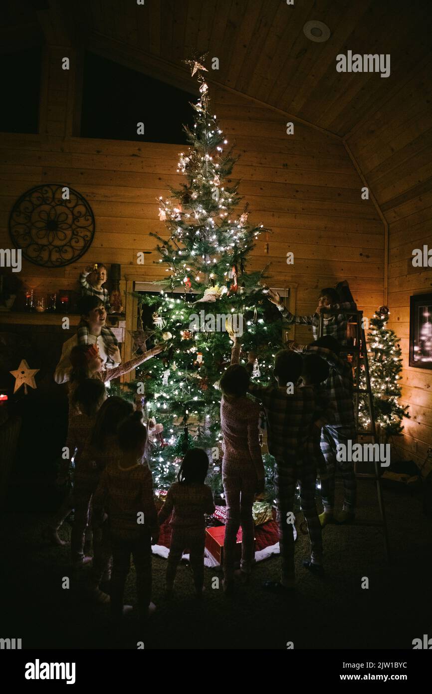 Kinder schmücken den weihnachtsbaum in der Hütte mit Verzierungen im Pyjama Stockfoto
