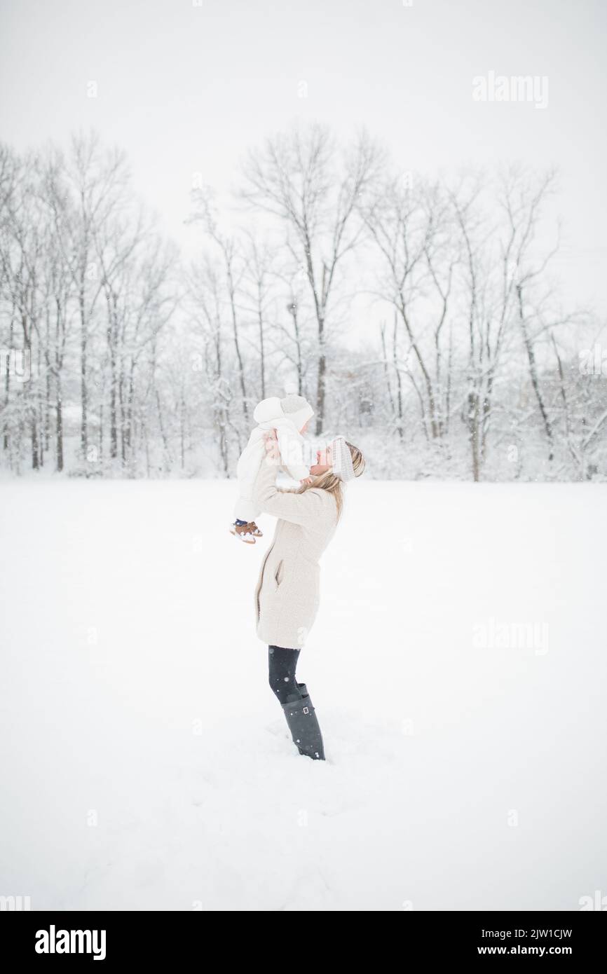 Die 30-jährige blonde kaukasische Mutter hält das Baby im Schnee hoch. Stockfoto