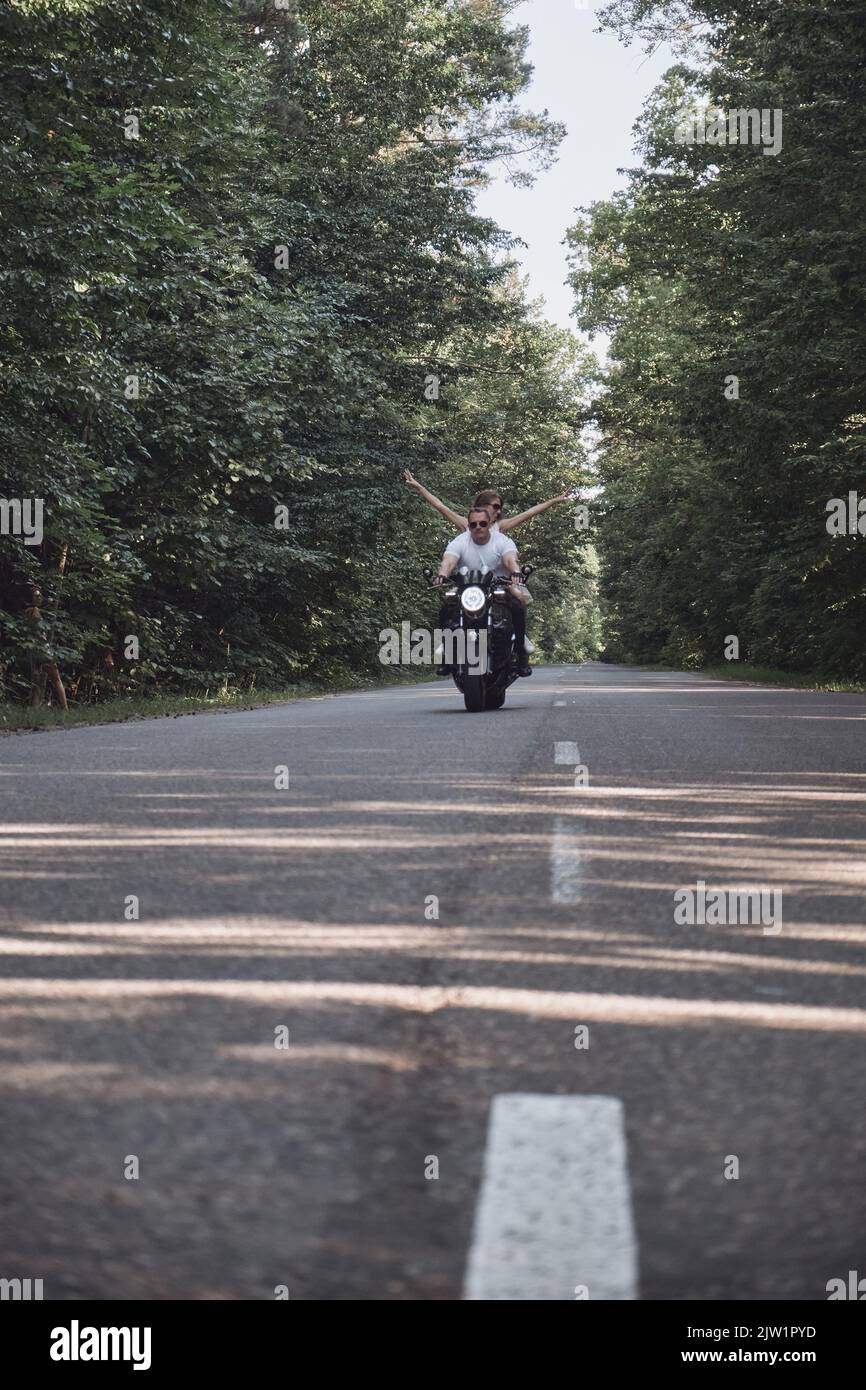 Ein junges glückliches Paar fährt mit dem Motorrad auf einer asphaltierten Straße im Wald, Freiheit und Geschwindigkeit Stockfoto