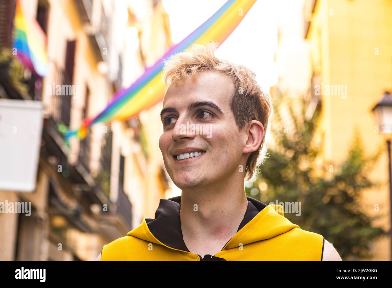 Von unten glücklicher nicht binärer Mann in stilvoller Kleidung lächelt und schaut weg auf verschwommenen Hintergrund von Häusern mit LGBT-Fahnen verziert Stockfoto