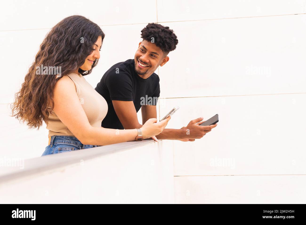 Lächelnder junger Mann, der auf dem Balkon auf die Freundin blickt und dabei auf dem Smartphone steht Stockfoto