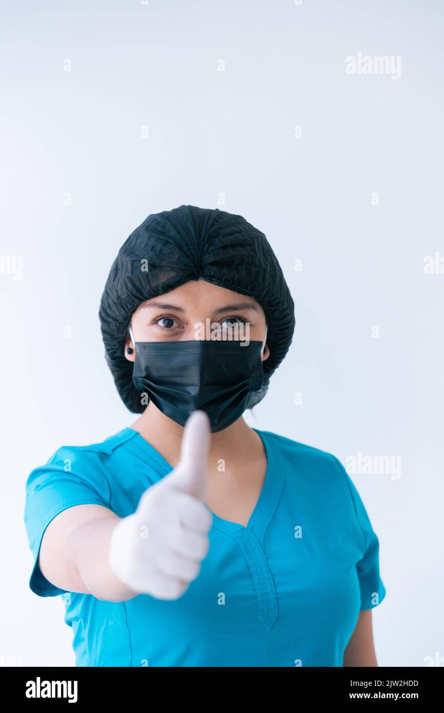 Inhalt junge weibliche Sanitäter in blau Scrubs Schutzhandschuhe Maske Kappe zeigt Daumen nach oben Geste und Blick auf die Kamera vor weißem Hintergrund Stockfoto