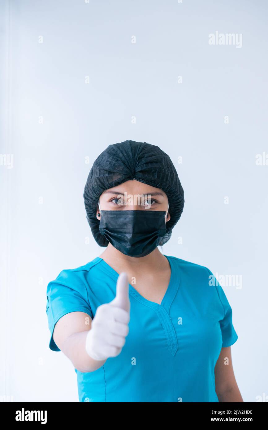 Inhalt junge weibliche Sanitäter in blau Scrubs Schutzhandschuhe Maske Kappe zeigt Daumen nach oben Geste und Blick auf die Kamera vor weißem Hintergrund Stockfoto
