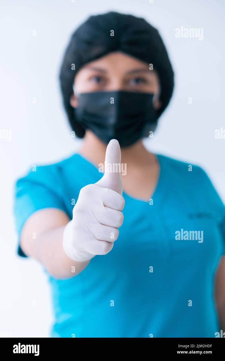 Inhalt junge weibliche Sanitäter in blau Scrubs Schutzhandschuhe Maske Kappe zeigt Daumen nach oben Geste und Blick auf die Kamera vor weißem Hintergrund Stockfoto
