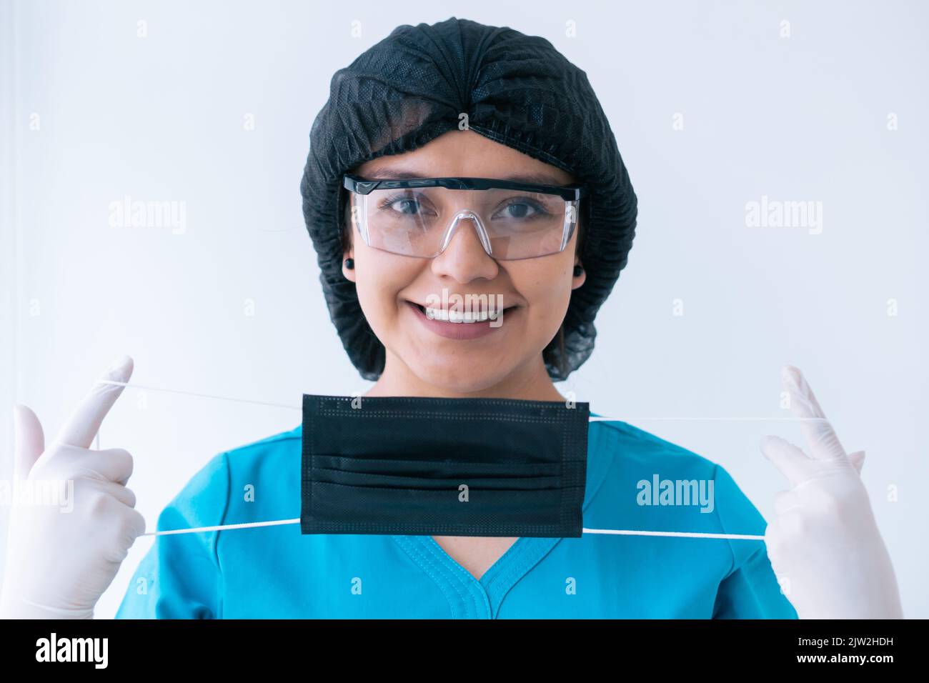 Fröhliche junge ethnische Ärztin in blauen Peelings Schutzhandschuhe Maskenkappe und Schutzbrille auf Schutzmaske setzen und lächeln Blick auf Kamera aga Stockfoto