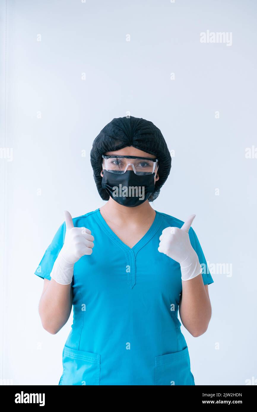 Inhalt junge weibliche Sanitäter in blau Scrubs Schutzhandschuhe Maskenkappe und Brille zeigt Daumen nach oben Geste und Blick auf Kamera gegen weißen Hintergro Stockfoto