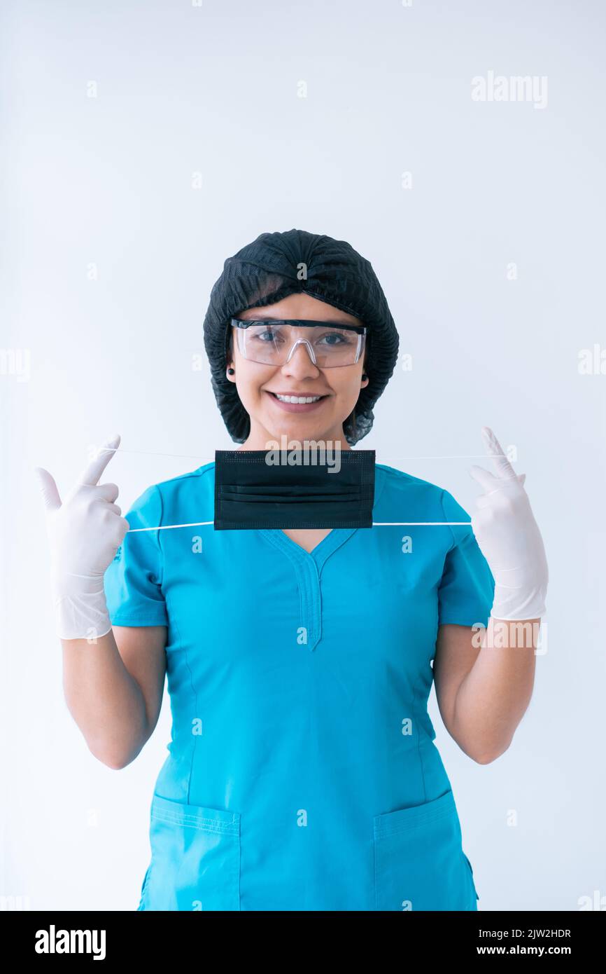 Fröhliche junge ethnische Ärztin in blauen Peelings Schutzhandschuhe Maskenkappe und Schutzbrille auf Schutzmaske setzen und lächeln Blick auf Kamera aga Stockfoto