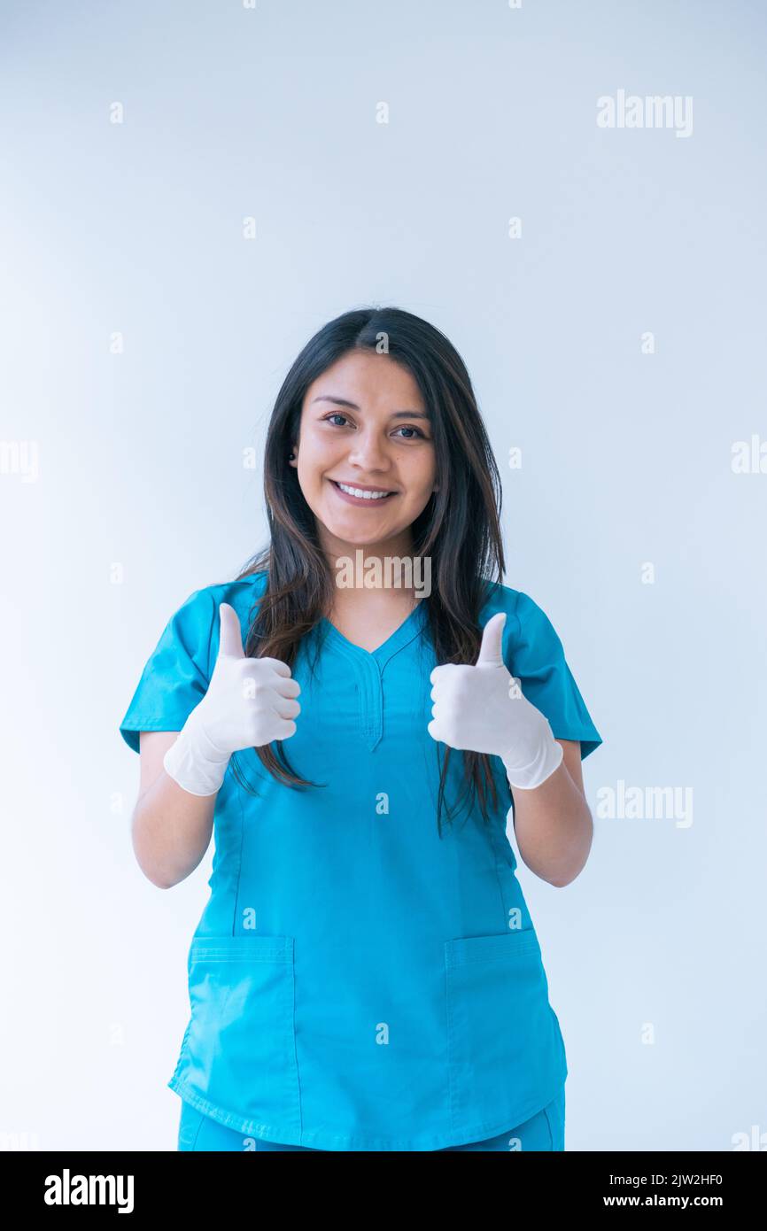 Entzückte junge ethnische Ärztin mit langen dunklen Haaren in blauen Peelings und Latexhandschuhen, die lächelten und die Daumen nach oben zeigten, während sie auf die Kamera schaute Stockfoto
