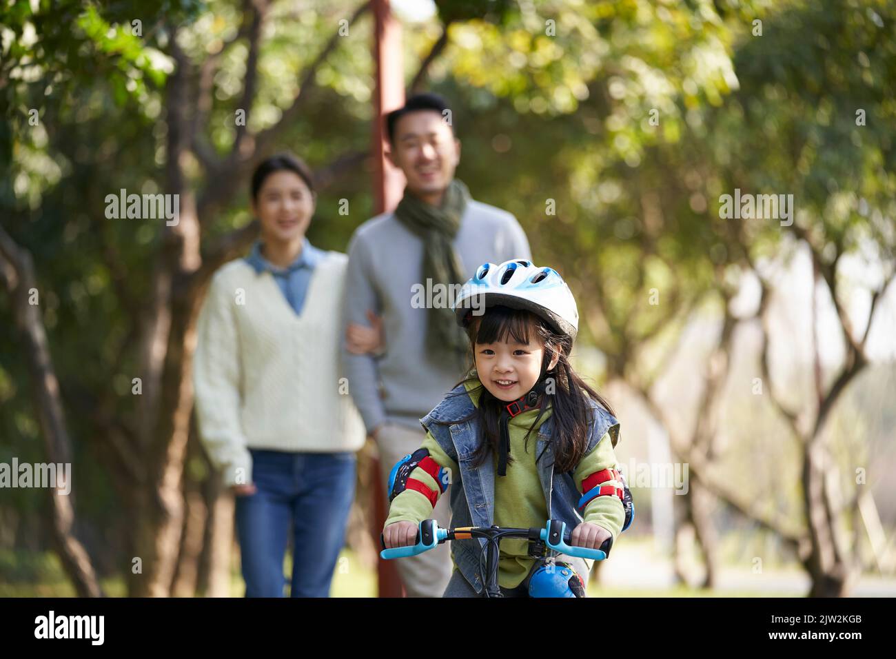 Kleine asiatische Mädchen Reiten Fahrrad im Stadtpark mit Eltern im Hintergrund Stockfoto