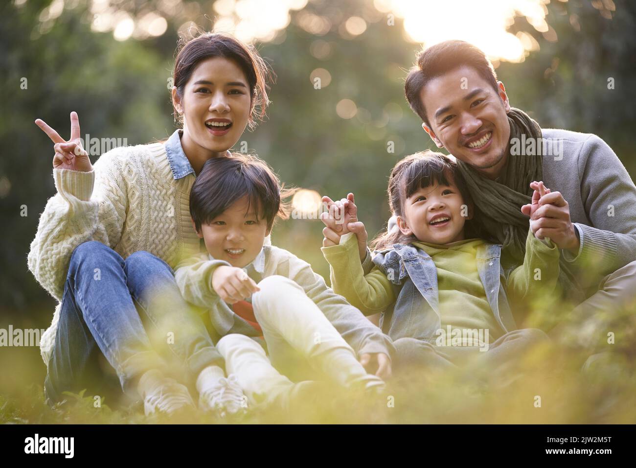 Outdoor-Porträt einer glücklichen jungen asiatischen Familie mit zwei Kindern auf Gras im Park sitzen Stockfoto