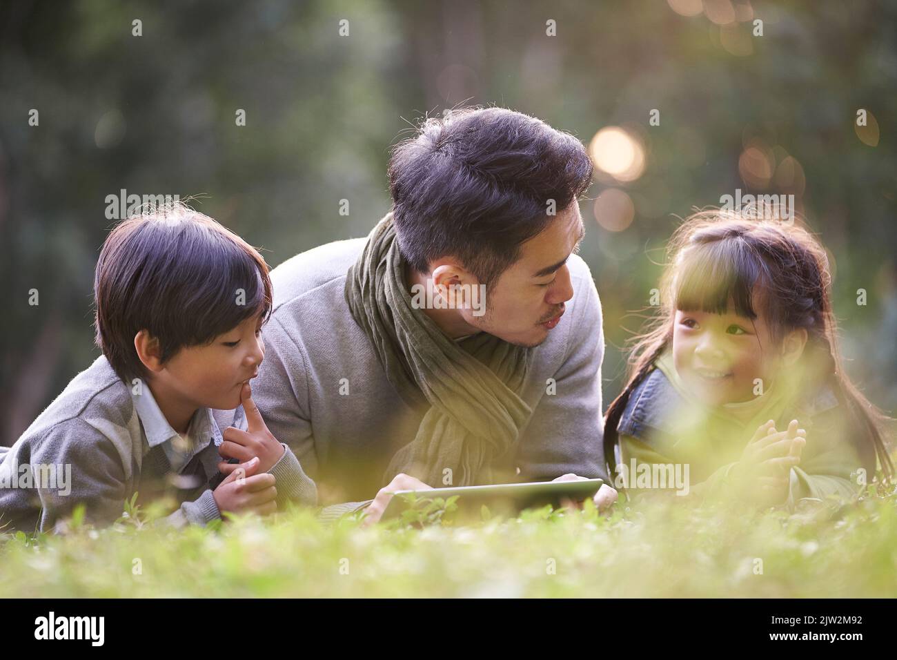 Junger asiatischer Vater, der auf Gras auf der Vorderseite liegt und Sohn und Tochter im Park erzählt Stockfoto