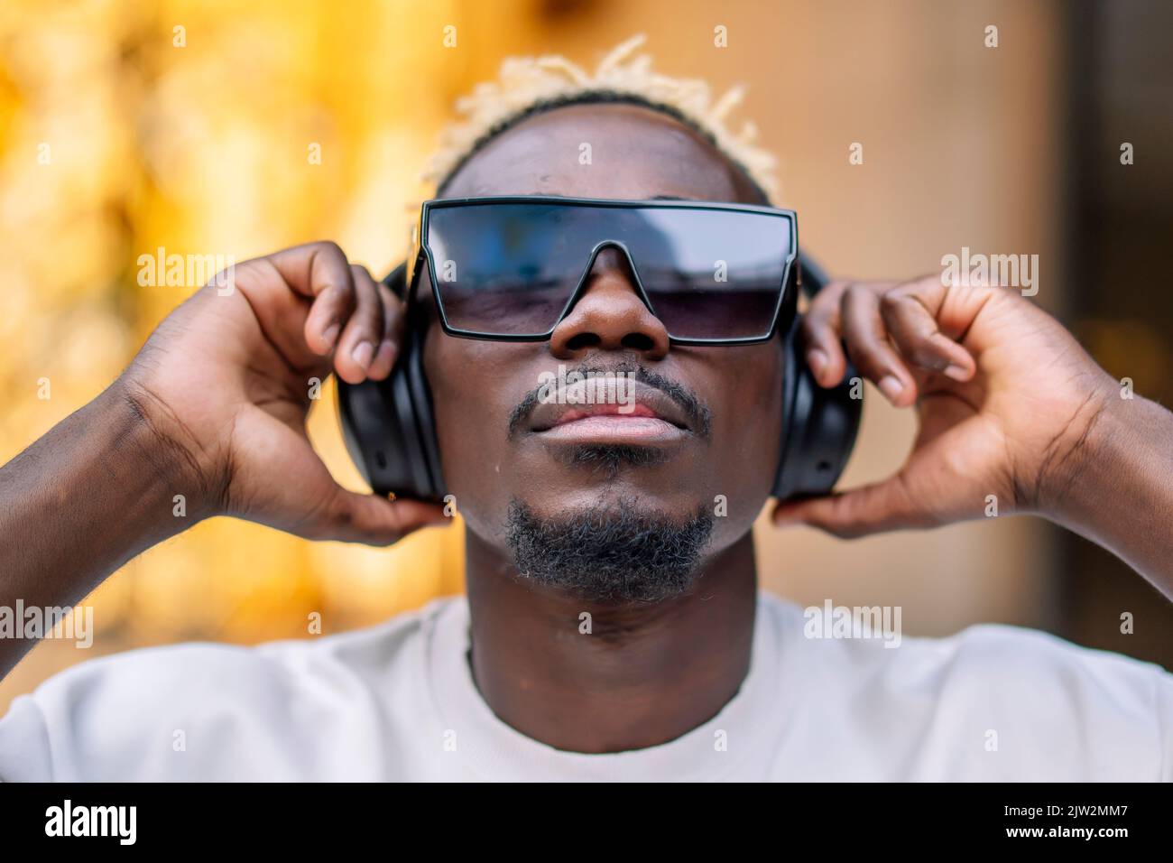 Seriöse afroamerikanische Meloman in T-Shirt und Sonnenbrille, die kabellose Kopfhörer anpasst und tagsüber in der Stadt gute Musik genießt Stockfoto