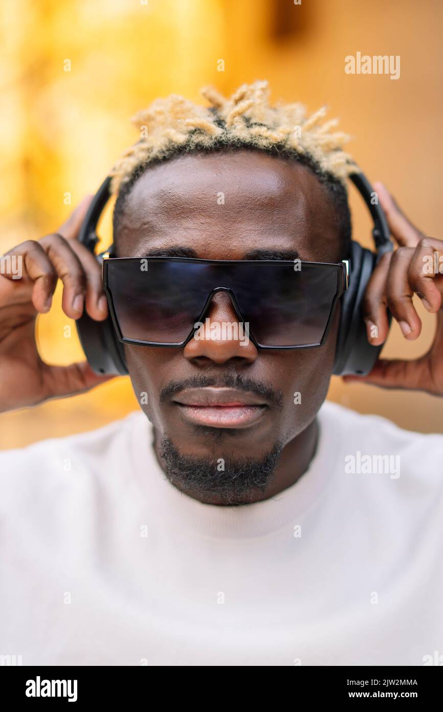 Seriöse afroamerikanische Meloman in T-Shirt und Sonnenbrille, die kabellose Kopfhörer anpasst und tagsüber in der Stadt gute Musik genießt Stockfoto