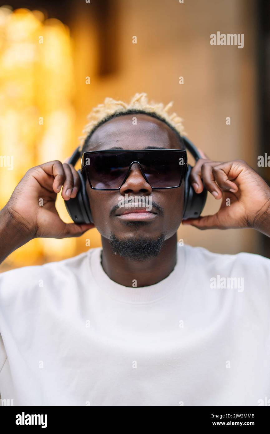 Seriöse afroamerikanische Meloman in T-Shirt und Sonnenbrille, die kabellose Kopfhörer anpasst und tagsüber in der Stadt gute Musik genießt Stockfoto