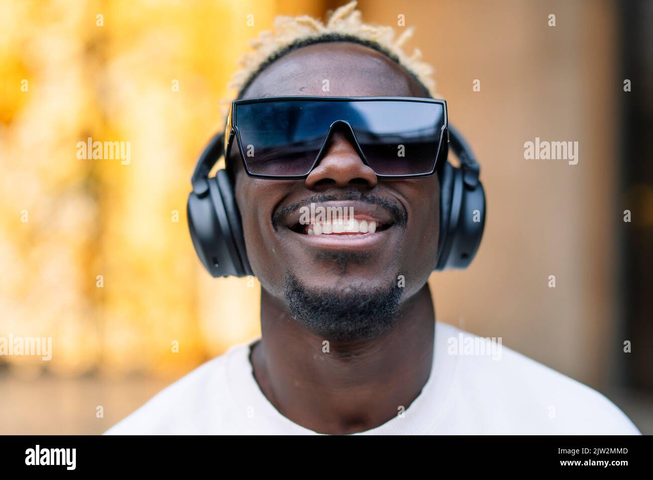 Fröhlicher afroamerikanischer Meloman in T-Shirt und Sonnenbrille, der kabellose Kopfhörer anpasst und lächelt, während er gute Musik mit geschlossenen Augen in d genießt Stockfoto