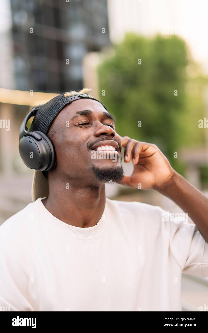 Fröhlicher afroamerikanischer Meloman in T-Shirt und Kappe, der kabellose Kopfhörer anpasst und lächelt, während er tagsüber gute Musik mit geschlossenen Augen genießt Stockfoto