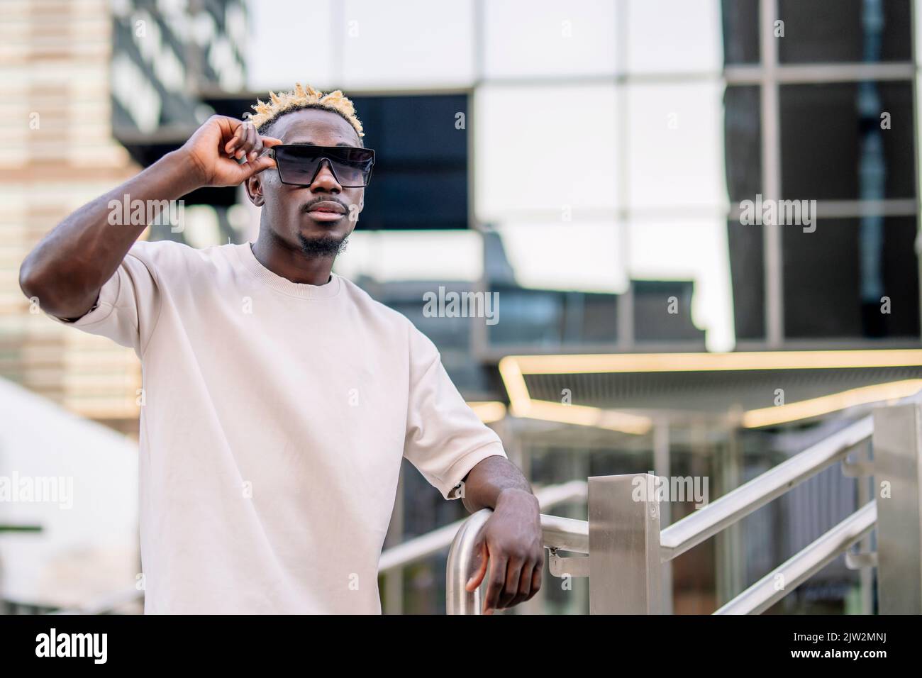 Junger afroamerikanischer Mann im lässigen T-Shirt mit gefärbtem Haar, das sich am Geländer stützt und tagsüber auf der modernen Stadtstraße eine Sonnenbrille anpasst Stockfoto