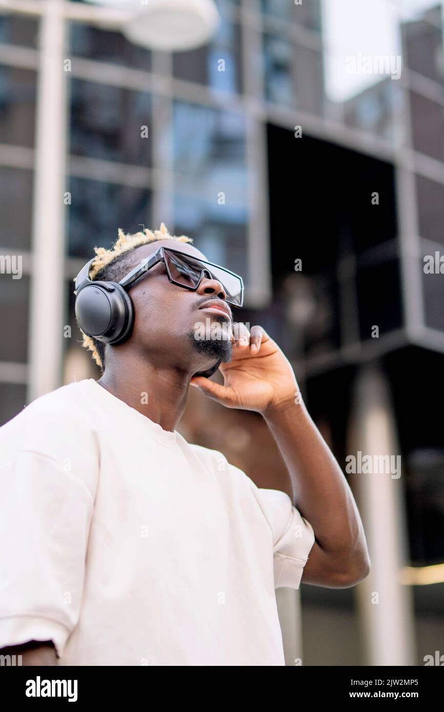 Von der unteren Seite sehen Sie einen ernsthaften afroamerikanischen Meloman in T-Shirt und Sonnenbrille, der kabellose Kopfhörer anpasst und dabei tagsüber gute Musik genießt Stockfoto