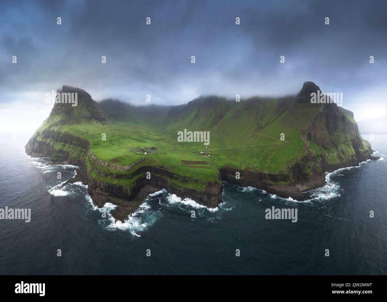 Atemberaubende Drohne Blick auf die Insel mit rauen felsigen Klippen mit grünem Gras bedeckt mit Siedlung Dorf umgeben von winkenden Meer gegen graue Klo Stockfoto