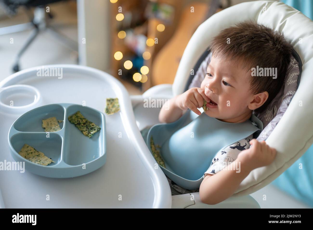 Kleiner Junge, der alleine auf seinem Hochstuhl zu Hause isst. Entzückendes, einjähriges Baby, das eine Mahlzeit in den Händen hält und zu Hause isst Stockfoto