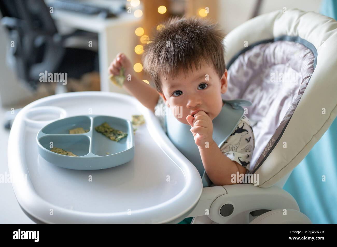 Kleiner Junge, der alleine auf seinem Hochstuhl zu Hause isst. Entzückendes, einjähriges Baby, das eine Mahlzeit in den Händen hält und zu Hause isst Stockfoto