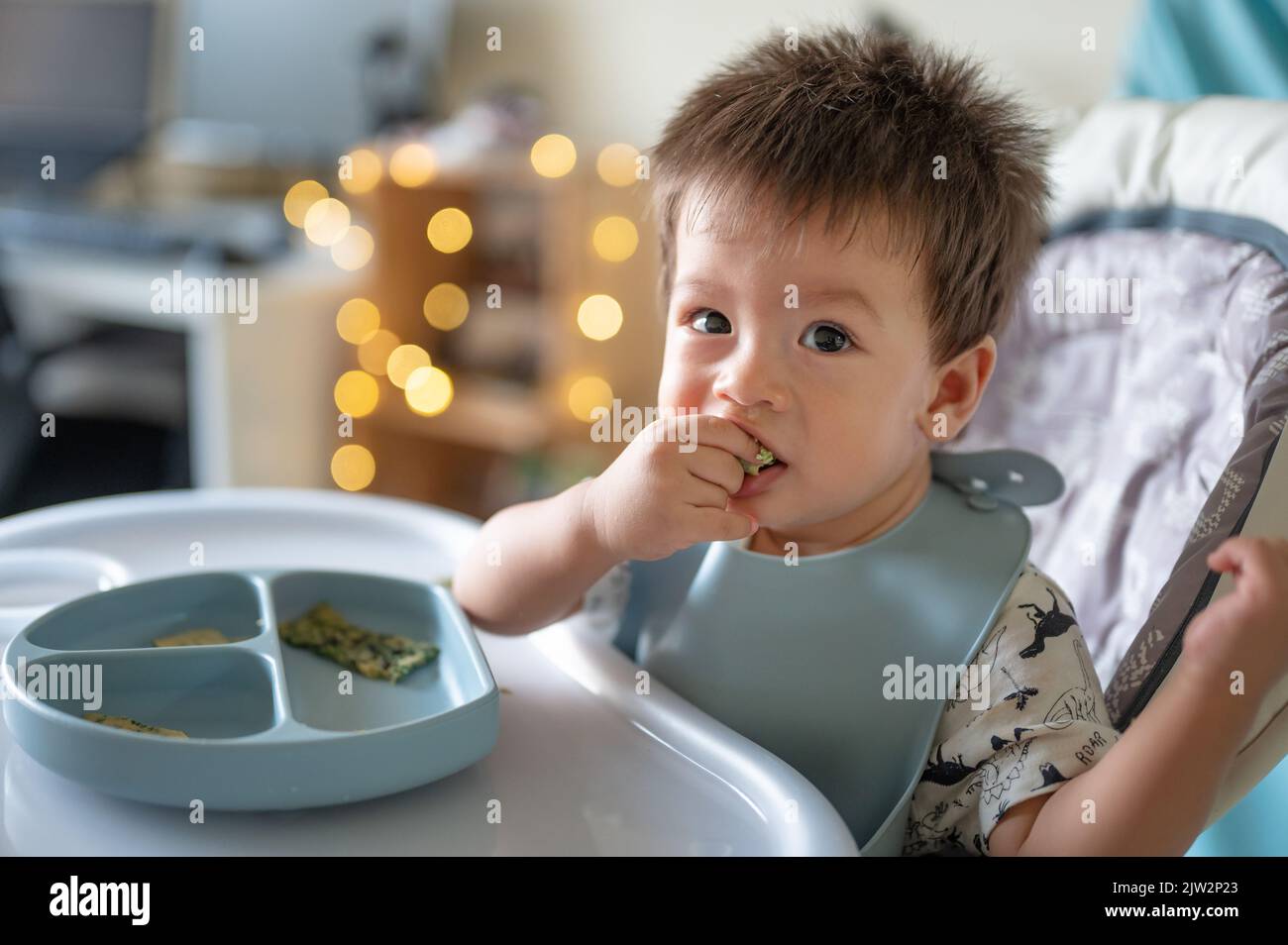 Kleiner Junge, der alleine auf seinem Hochstuhl zu Hause isst. Entzückendes, einjähriges Baby, das eine Mahlzeit in den Händen hält und zu Hause isst Stockfoto