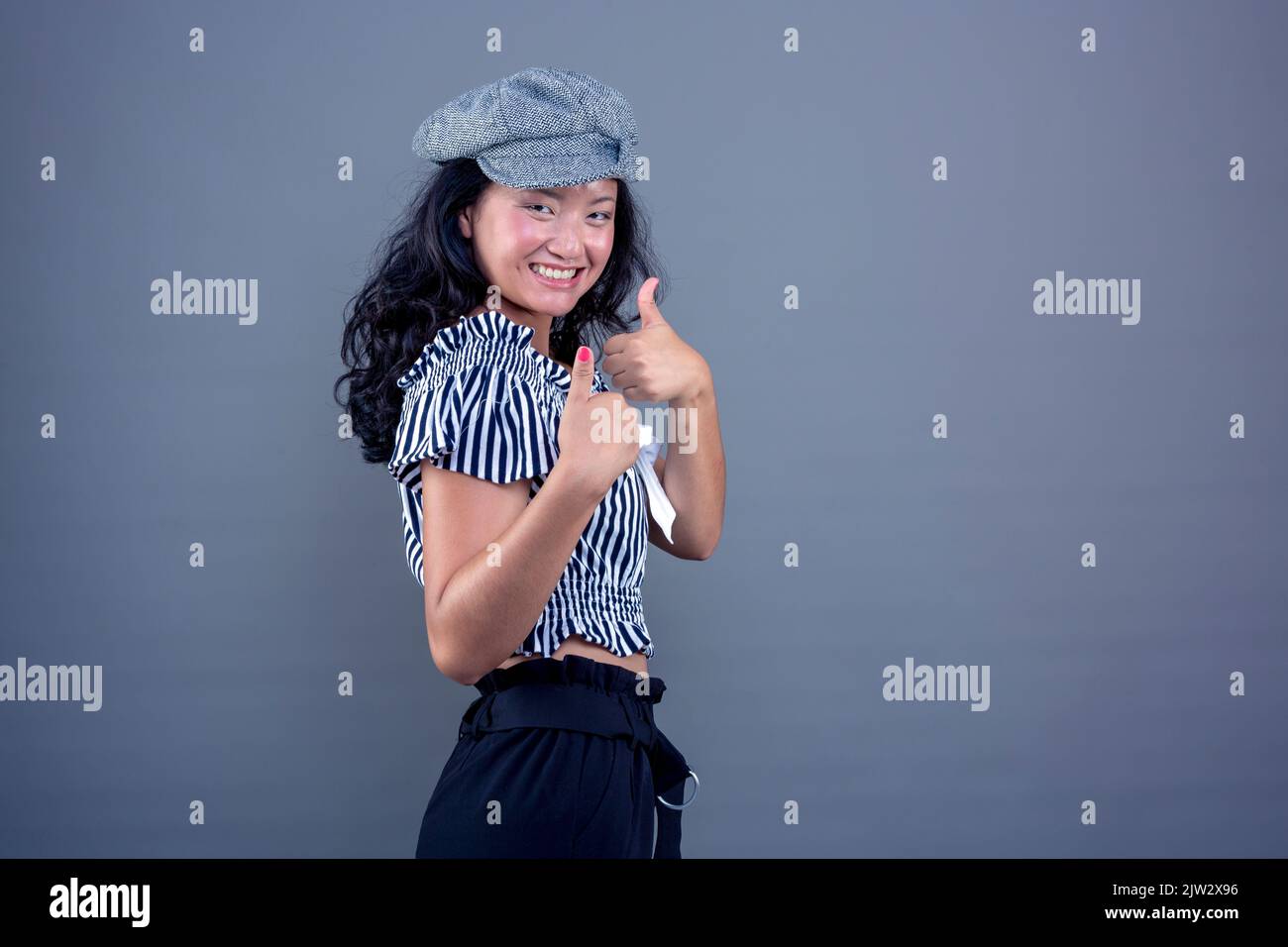 Nette asiatische ethnische Frau, die Daumen nach oben gibt, als Zeichen eines OK lächelnd und isoliert auf grauem Hintergrund Stockfoto