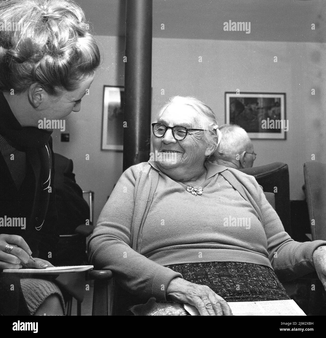 1965, historisch, eine ältere Dame, die auf einem Stuhl im Loungebereich eines Altersheims sitzt, von einer Journalistin interviewt wird, um vielleicht ihre Lebensgeschichte zu erzählen. Stockfoto