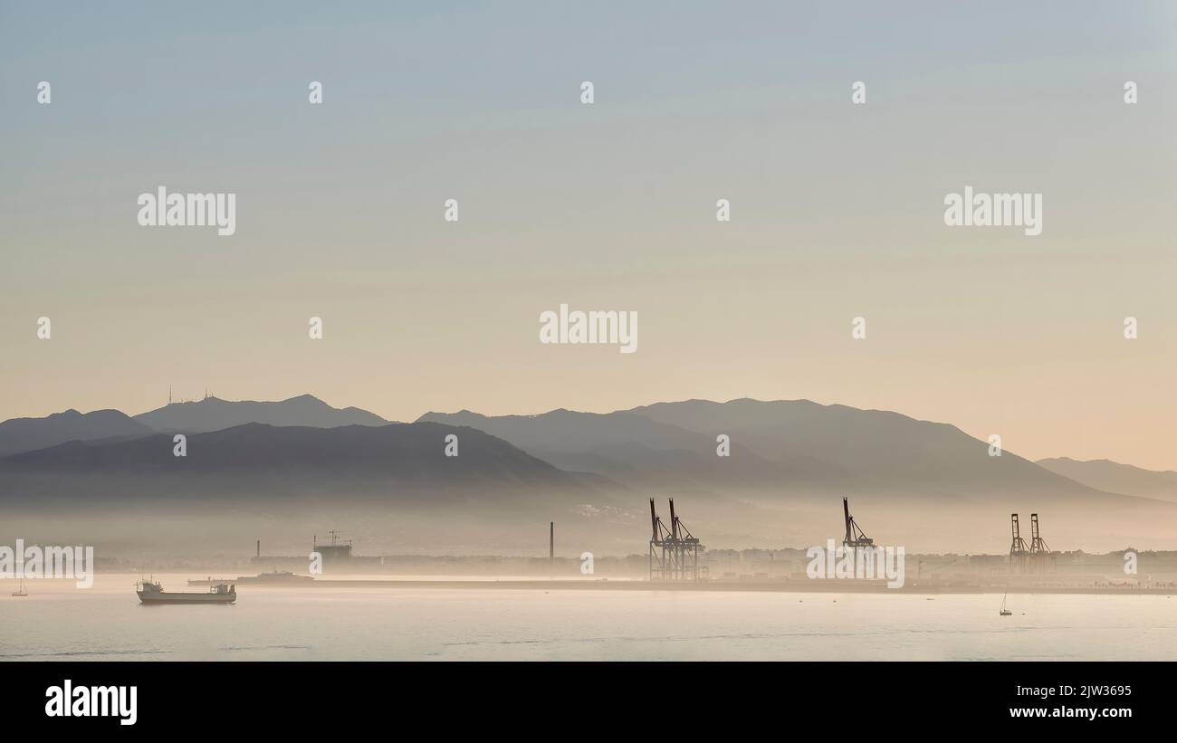 Sonnenuntergang im Hafen von Malaga mit Nebel durch Feuchtigkeit mit Kräne, Schiffen und Bergen dahinter Stockfoto