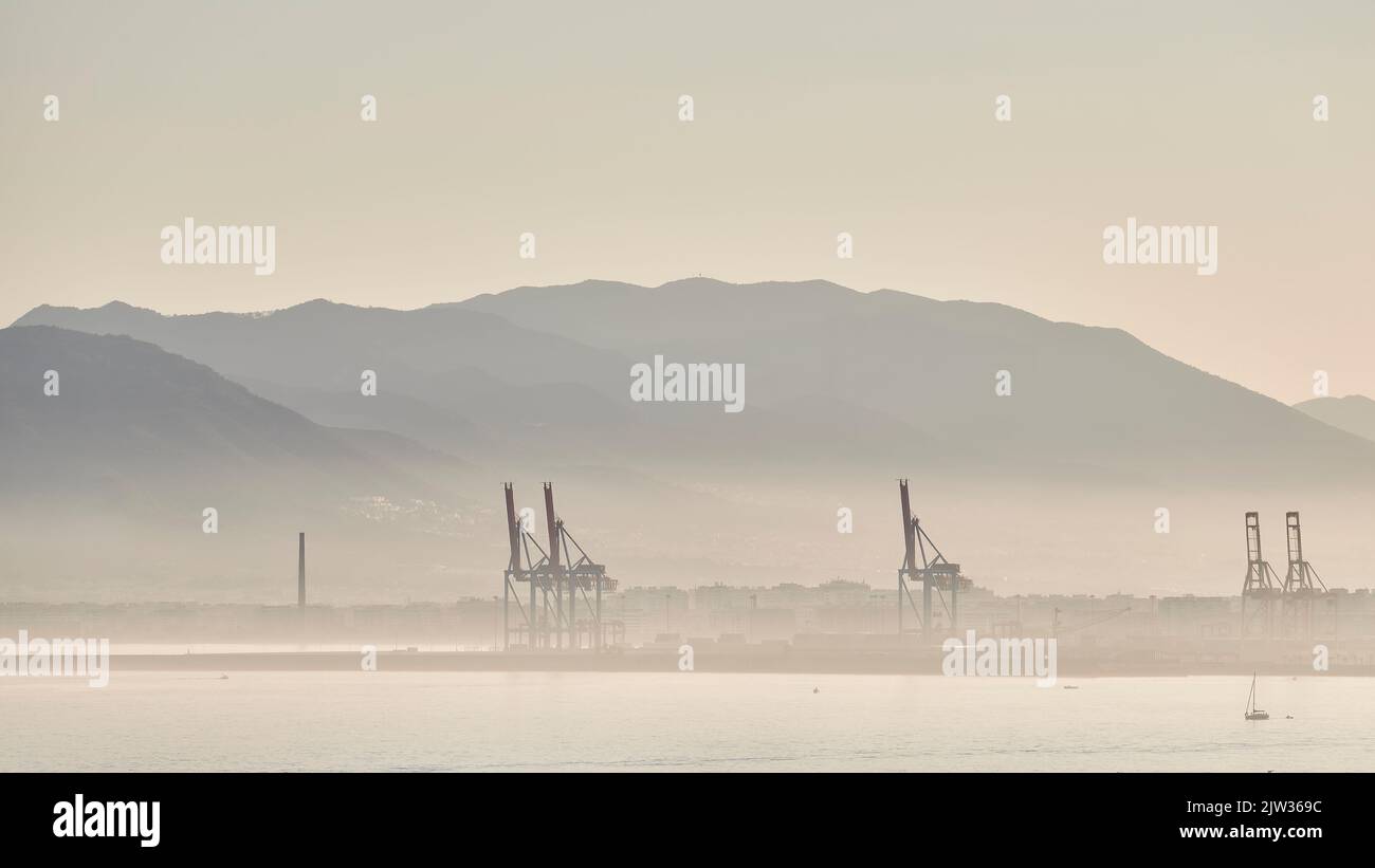 Sonnenuntergang im Hafen von Malaga mit Nebel durch Feuchtigkeit mit Kräne, Schiffen und Bergen dahinter Stockfoto