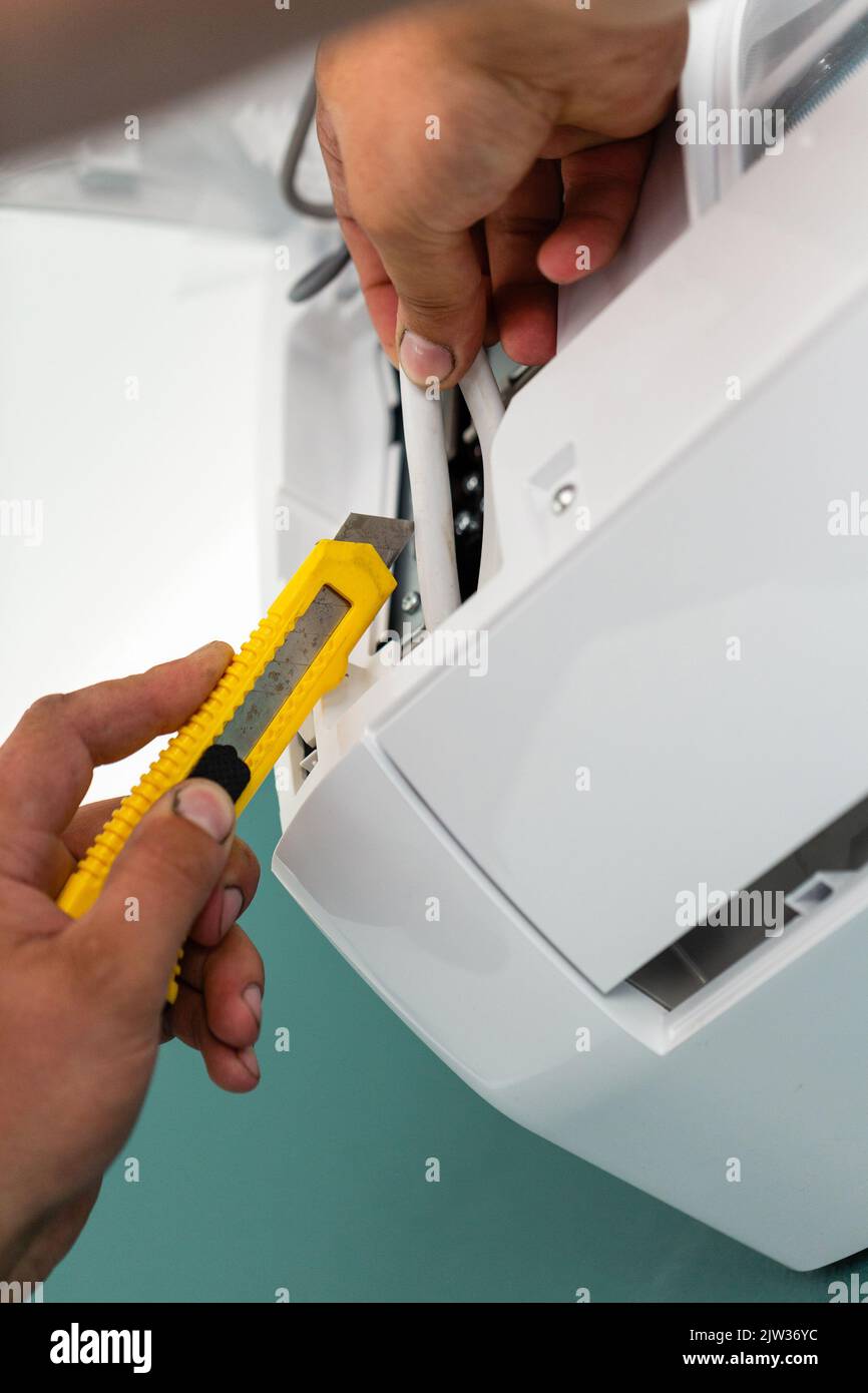 Elektroinstallierer mit Schreibwarenmesser schneidet und streift Drähte, während die Klimaanlage installiert wird. Nahaufnahme eines gelben Schreibwarenmessers in den Händen des Profess Stockfoto