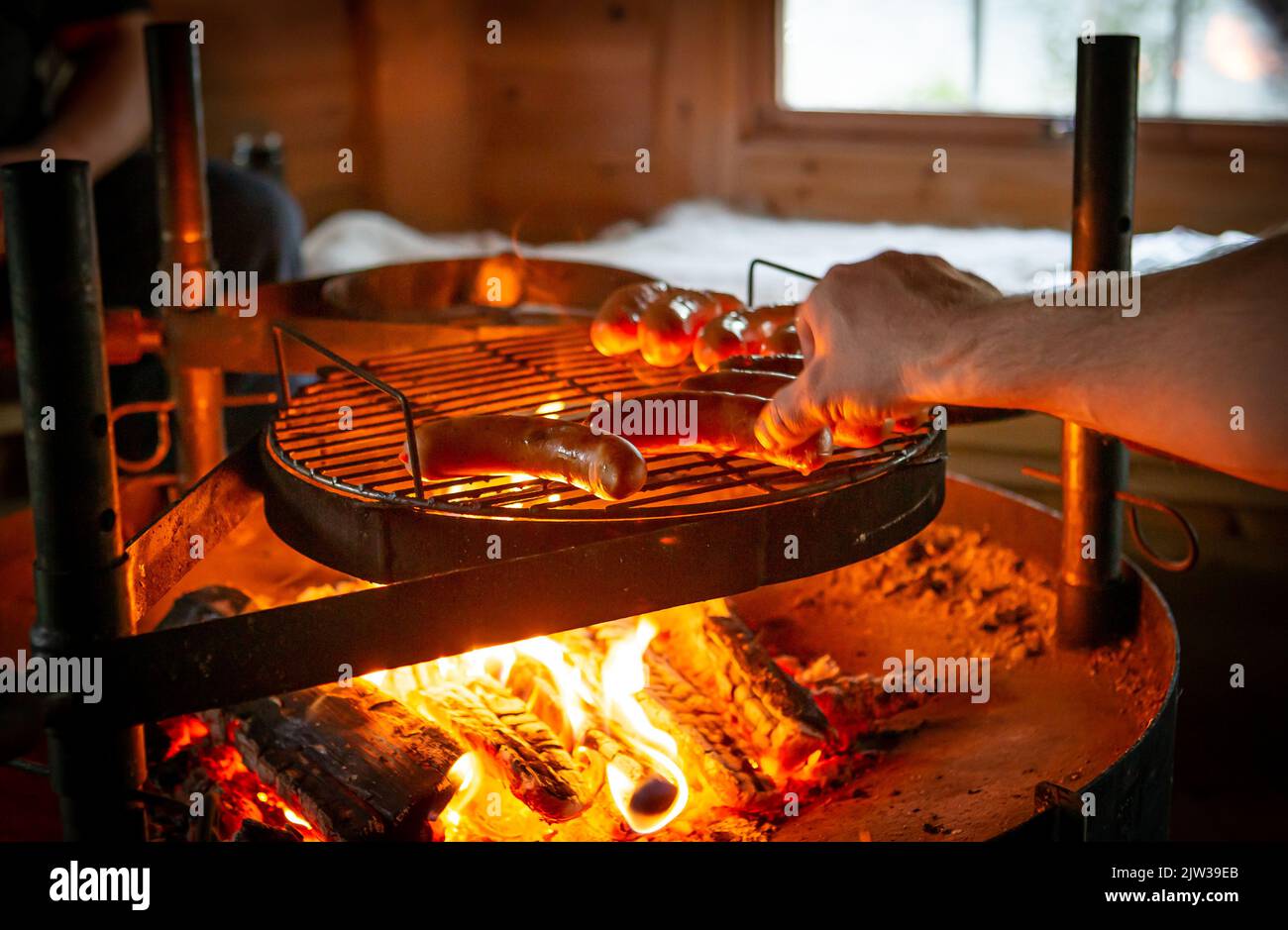 Kochen auf Feuer. Würste werden auf einem offenen Feuer in einem Grill gebraten. Würste von Hand umdrehen. Im Hintergrund befindet sich eine Wand des hölzernen Grillschuppen. Stockfoto