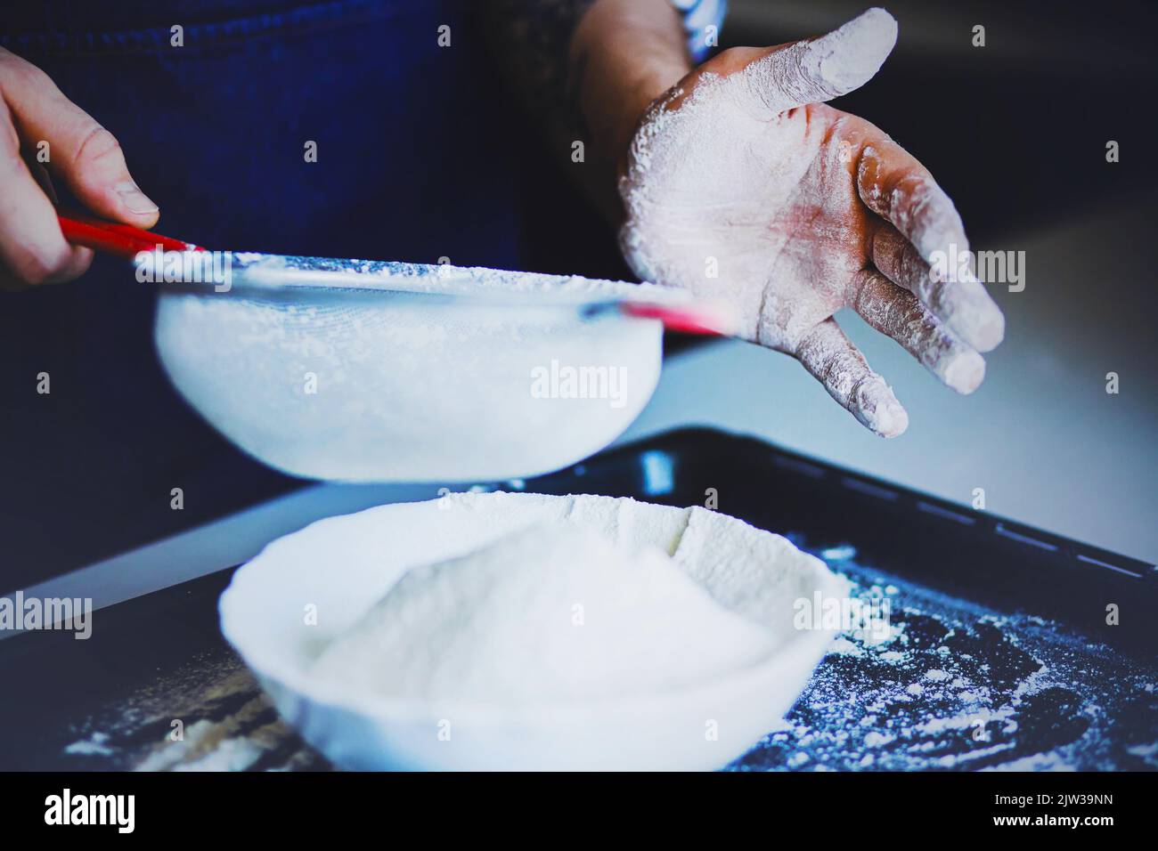 Ein Mann in einer blauen Schürze sieben Mehl durch ein Sieb in einen weißen Teller. Der Prozess des Kochens zu Hause. Mehl und Gebäck. Stockfoto