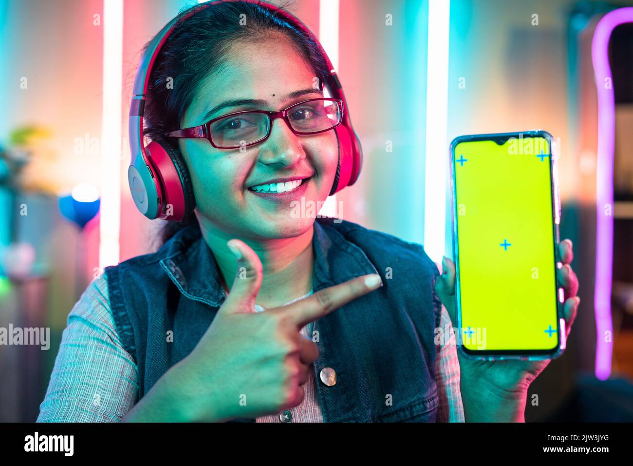 Lächelnde Frau mit schnurlosem Headset, das ein Mobiltelefon mit grünem Bildschirm zeigt, indem sie mit dem Finger zeigt, während sie die Kamera zu Hause betrachtet – Spielkonzept Stockfoto