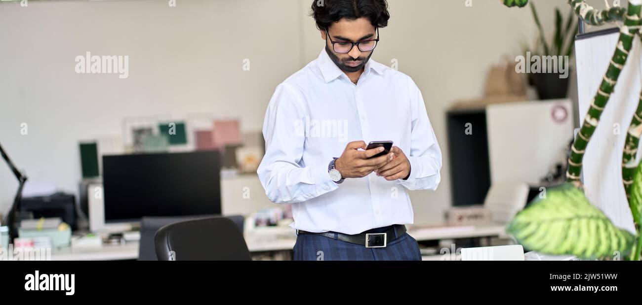 Eleganter junger indischer Geschäftsmann, der im Büro Smartphone verwendet. Stockfoto