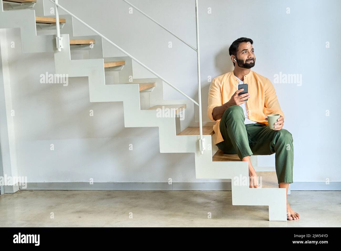 Indischer Mann, der zu Hause auf einer Treppe sitzt und Ohrstöpsel mit einem Mobiltelefon trägt. Stockfoto