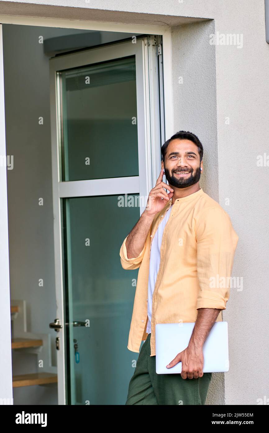 Lächelnder indischer Geschäftsmann, der vor dem Büro steht und mit Ohrhörern telefoniert. Stockfoto